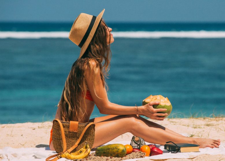 Mulher sentada na areia da praia, usando chapéu e biquíni, segurando água de coco, em cenário de verão com mar ao fundo, representando cuidados conscientes com a pele e proteção solar no verão.